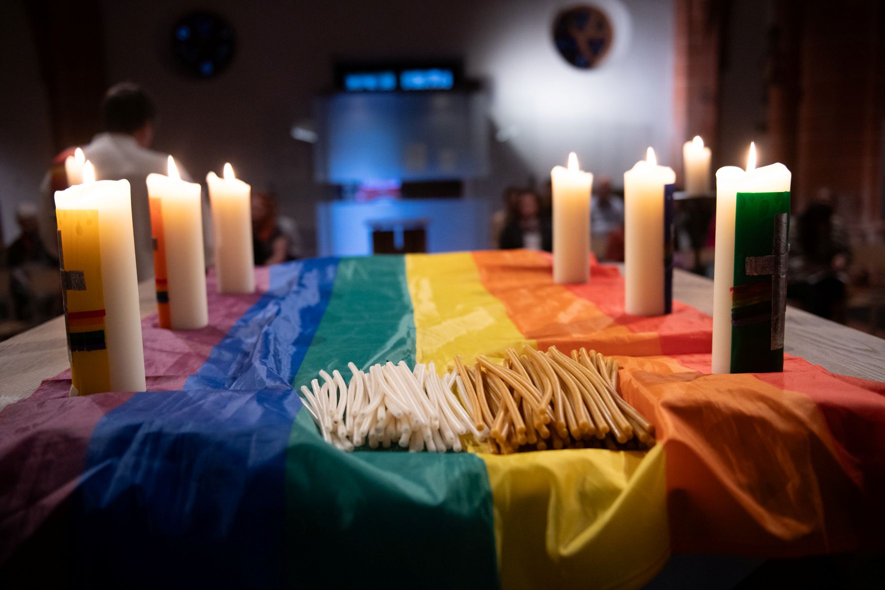 SEchs brennende Kerzen stehen auf einem Altar, der mit einem regenbogenfarbenen Tuch bedeckt ist.