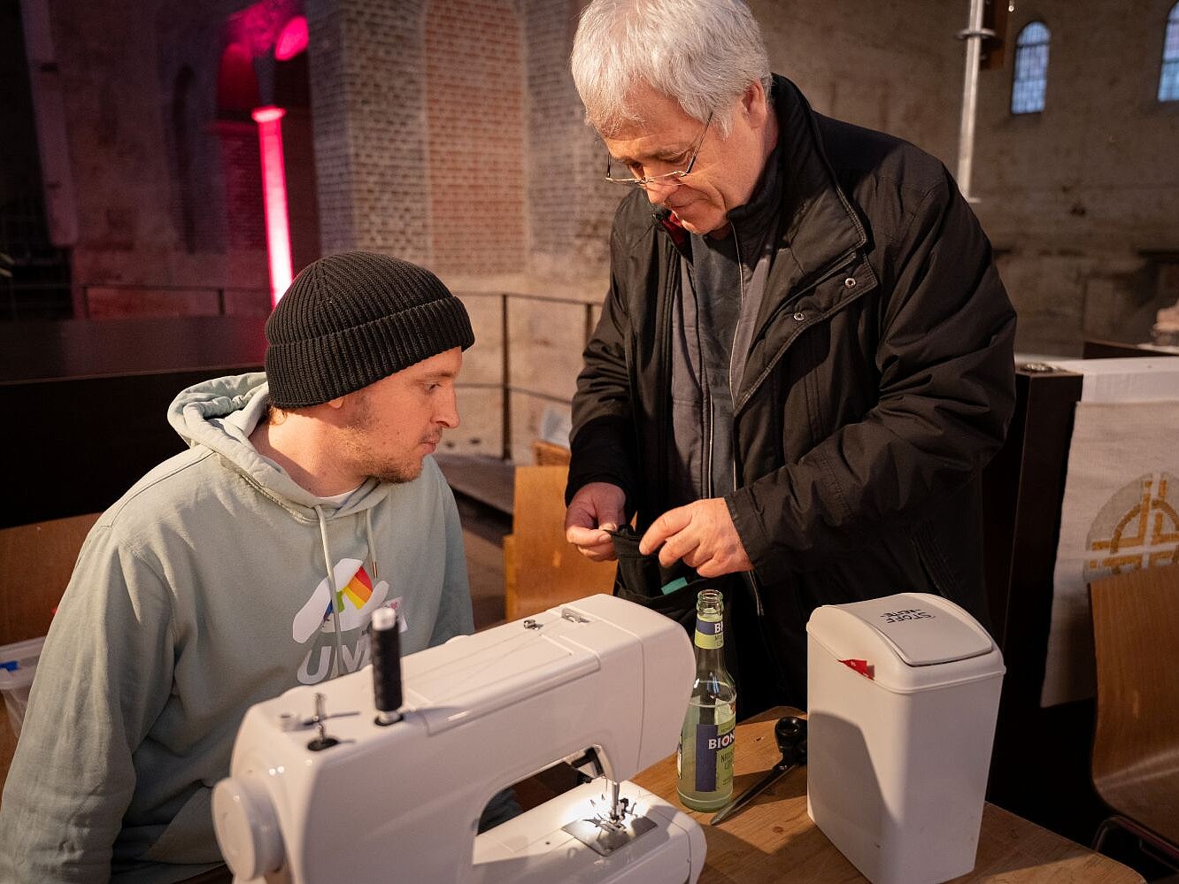 Ein junger Mann mit Kapuzenpulli und Mütze sitzt hinter einer Nähmaschine und erklärt einem älteren Mann, der neben ihm steht etwas.