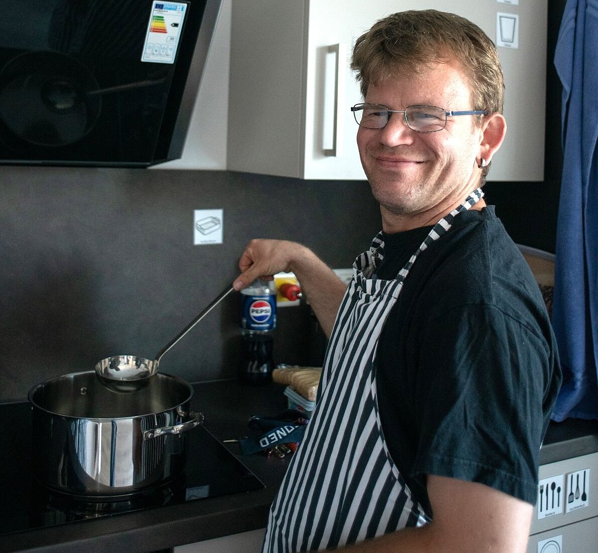 Ein junger Mann mit Brille trägt eine gestreifte Kochschürze, hält eine Kelle in der Hand und rührt in einem Topf.