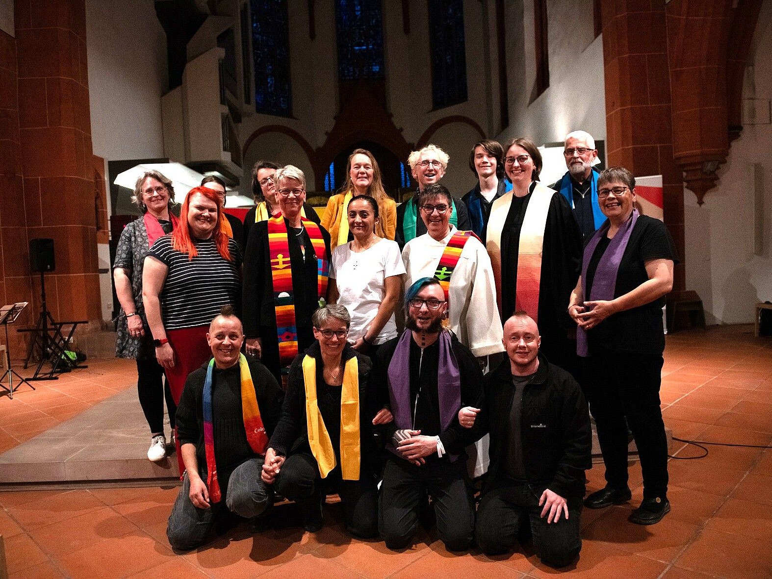 Eine Gruppe unterschiedlichster Menschen, viele mit regenbogenfarbenen Stolas stehen und sitzen als Gruppe in einer Kirche. 