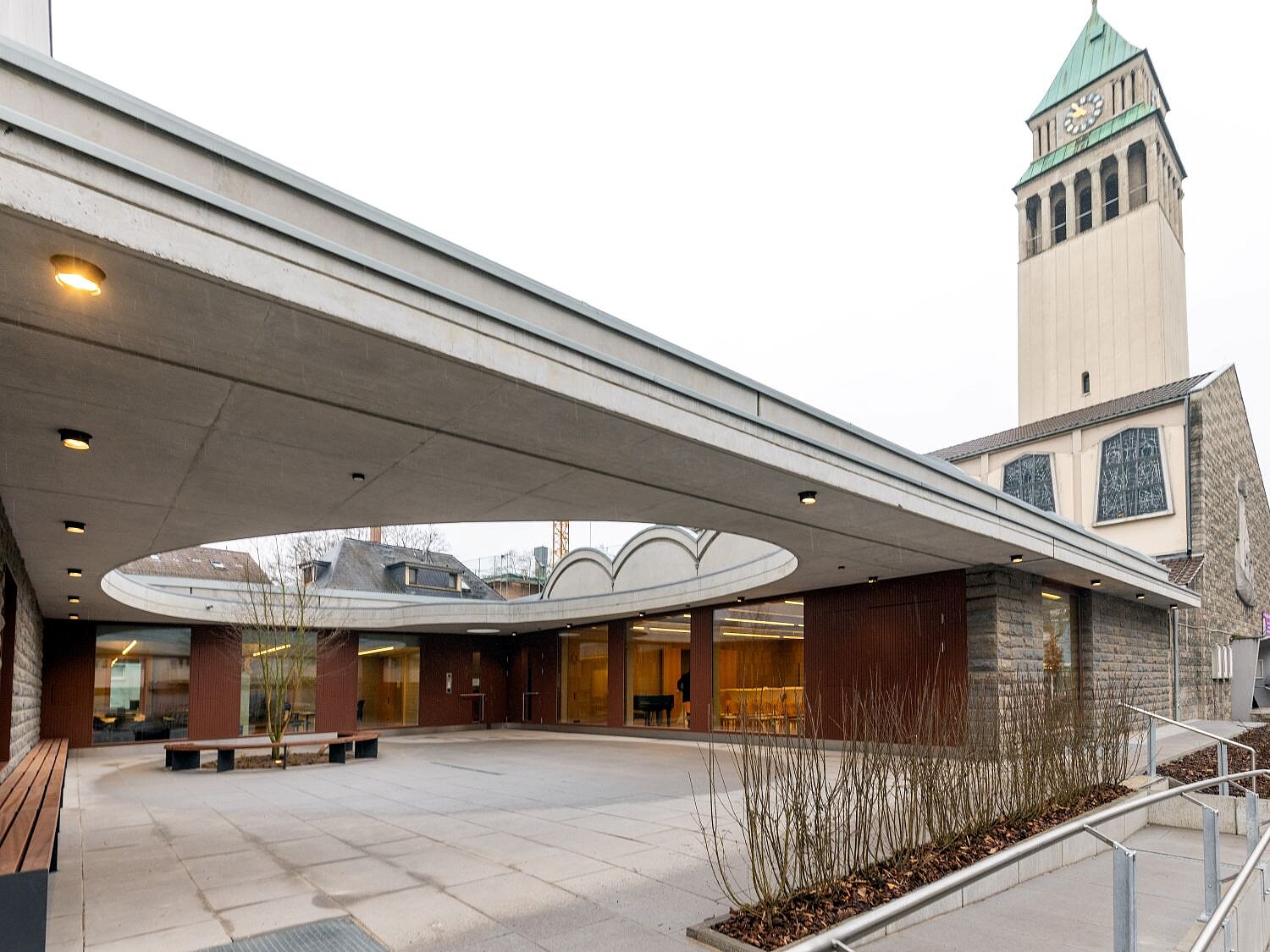 Eine weiße Kirche mit grünem Dach, daran schließt sich ein überdachter Platz an, der in der Mitte eine runde Öffnung hat.