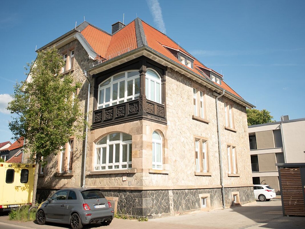 Ein älteres Eckhaus mit rotem Dach, davor parken zwei Autos.