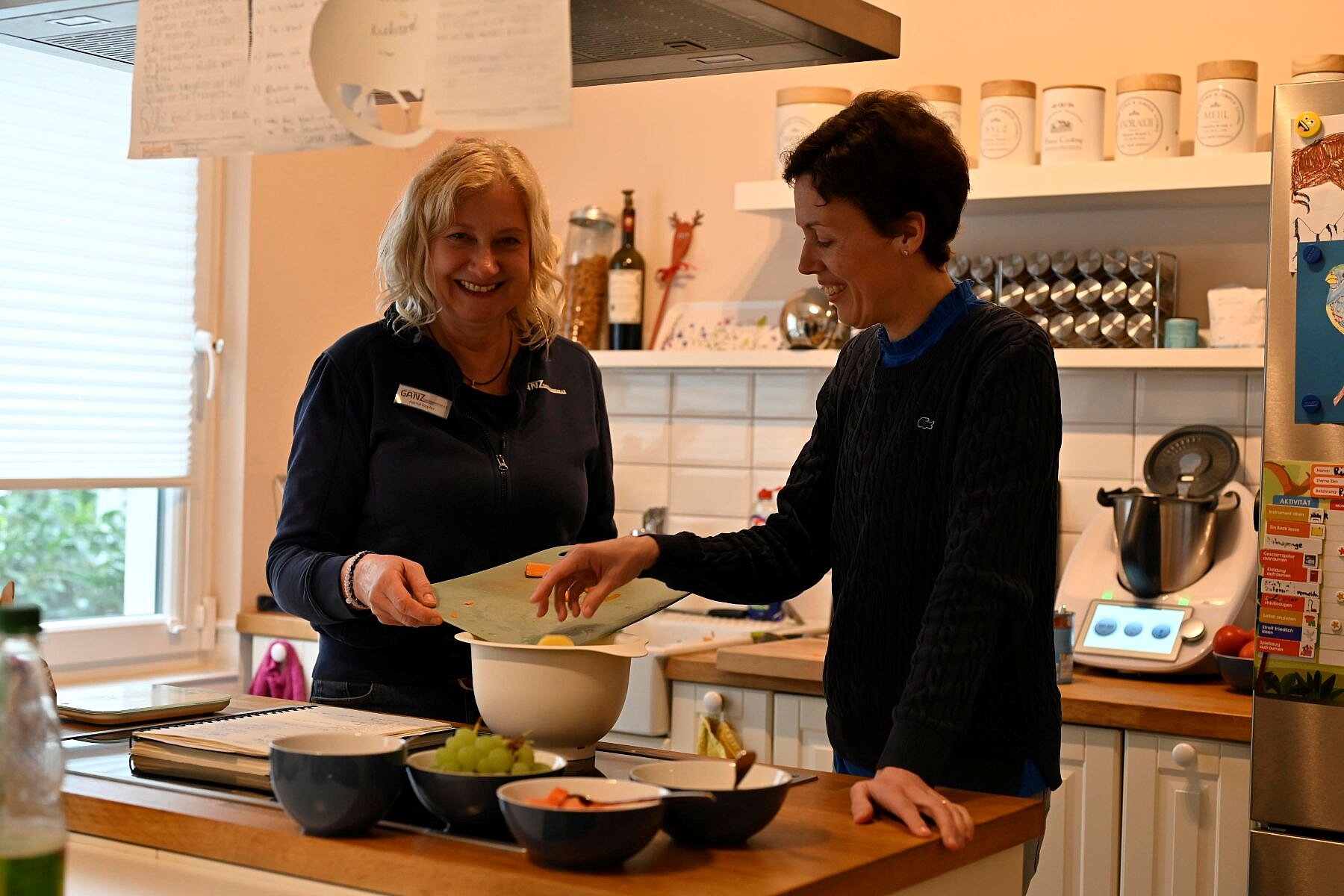 Zwei Frauen stehen in der Küche und kochen, sie haben gerade Gemüse geschnitten, dass sie eine Schüssel füllen.