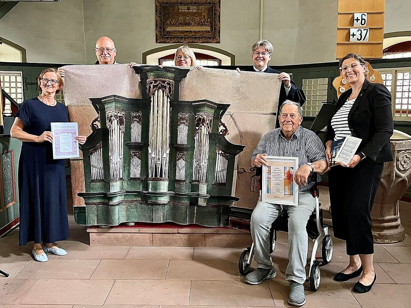 Im Kirchenraum stehen fünf Menschen und ein Mann sitzt auf dem Rollator. Drei Stehende halten ein großes Bild fest auf dem eine Orgel abgebildet ist