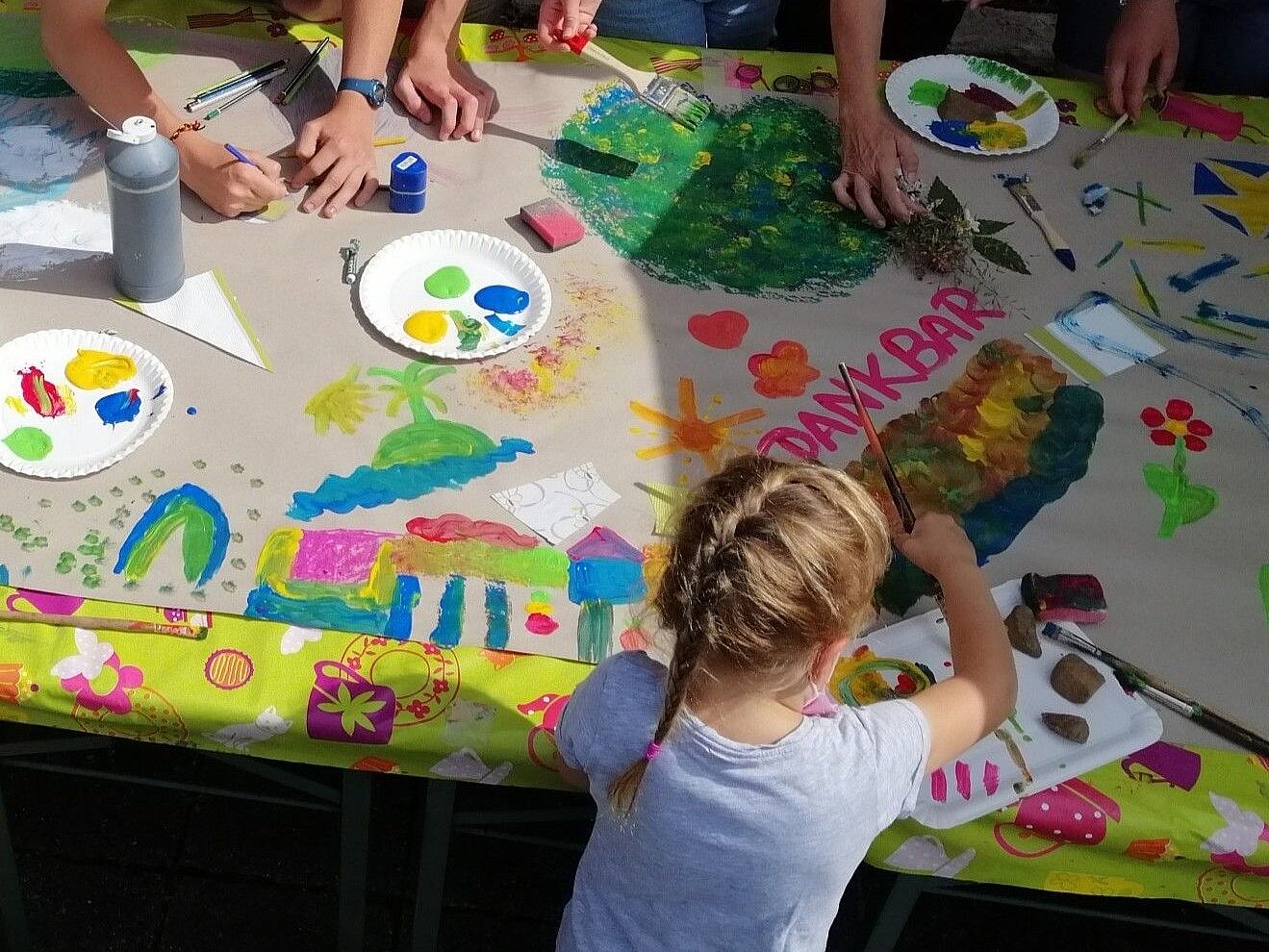 Kinder bemalen eine große Pappe, die auf einem Tisch liegt mit verschiedenen Farben.