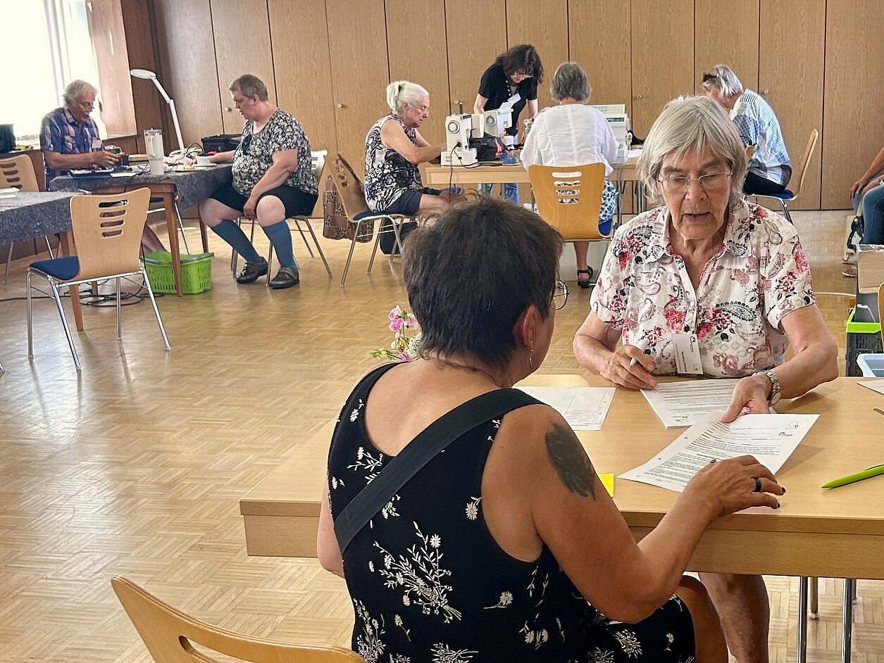 Zwei Frauen sitzen an einem Tisch und füllen Formulare aus im Hintergrund sind mehrere Tische und Menschen zu sehen an denen Dinge repaiert werden.