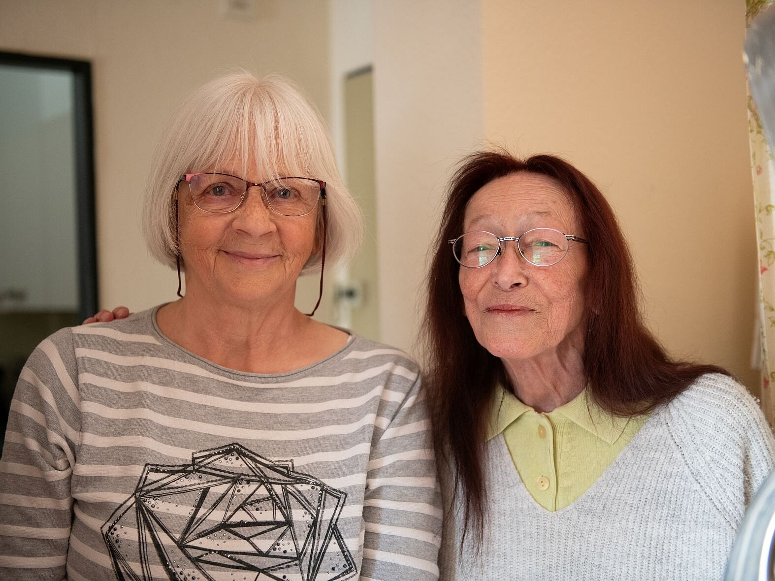 Zwei ältere Frauen, eine mit weißem Haar und gestreiftem Pulli und eine mit braunen längeren Haaren, stehen nebeneinander und lächeln.