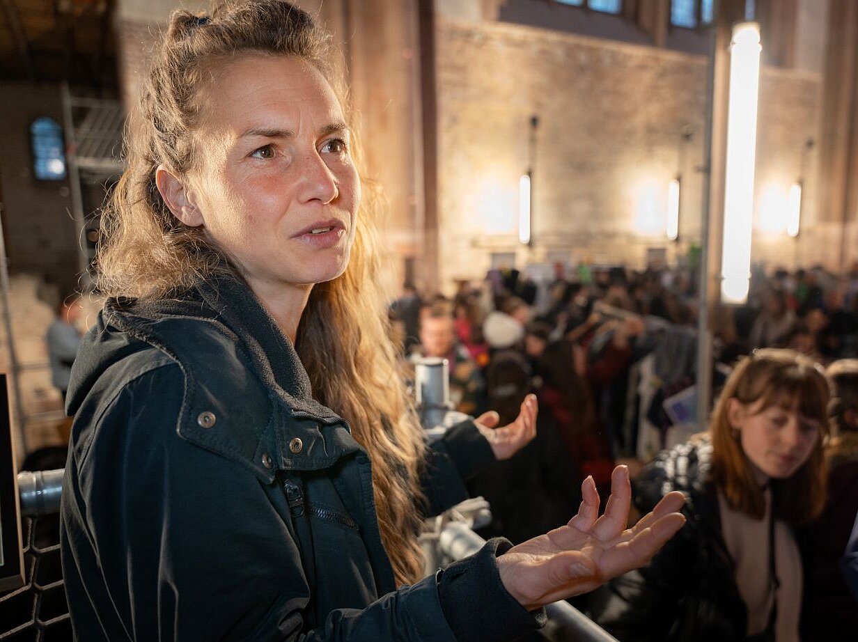 Eine Frau mit braunen lockigen Haaren steht in einer Kirche und spricht.