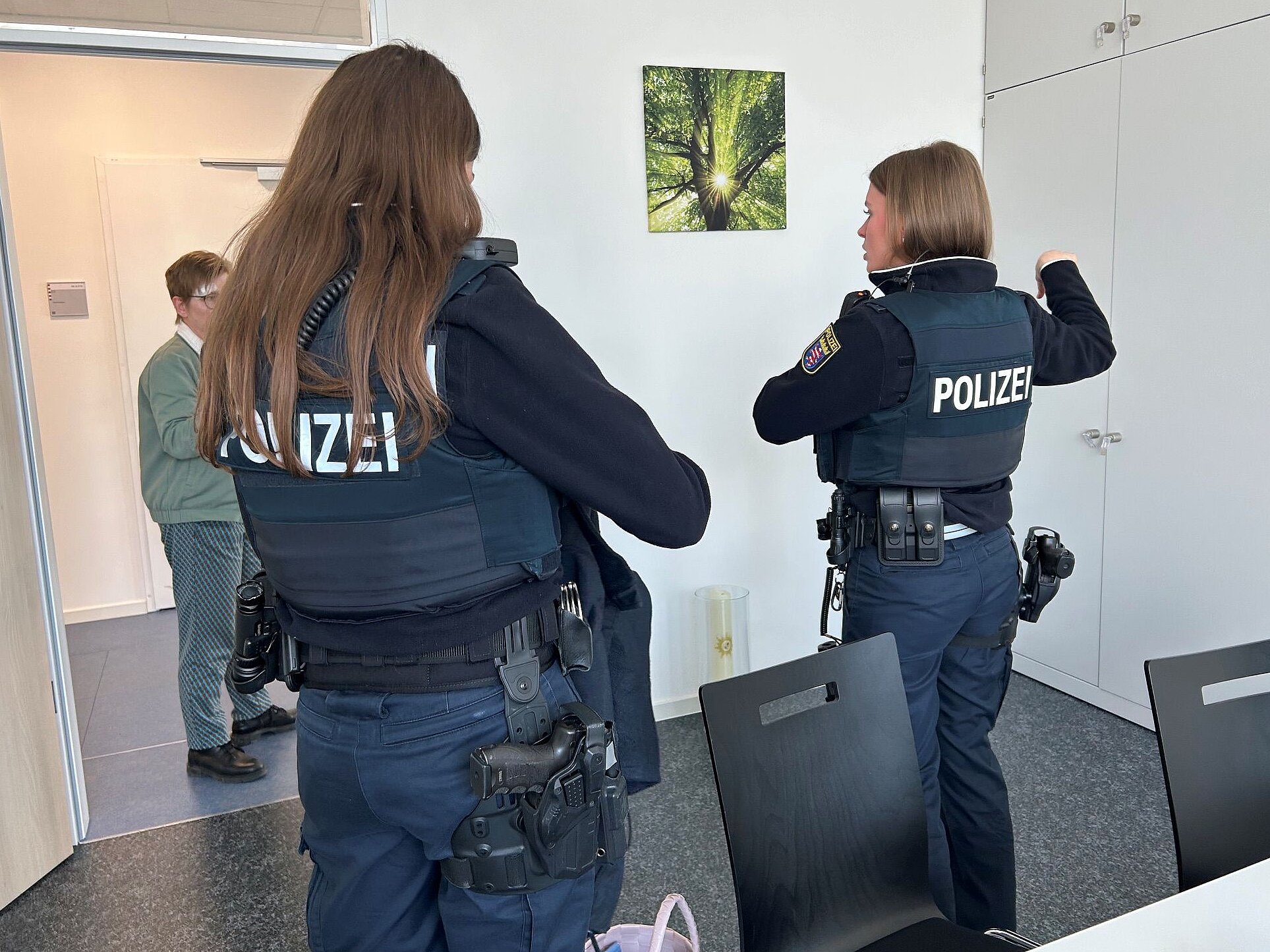 Zwei Polizistinnen in Uniform sprchen mit einer Frau in grüner Jacke in einem Büroraum.