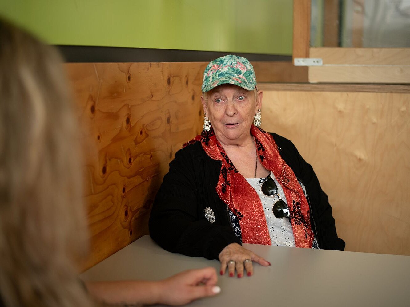 Eine Frau mit einer grünen Kappe und einem roten Schal sitzt am Tisch und unterhält sich mit einer anderen Frau von der man nur die Haare sieht.